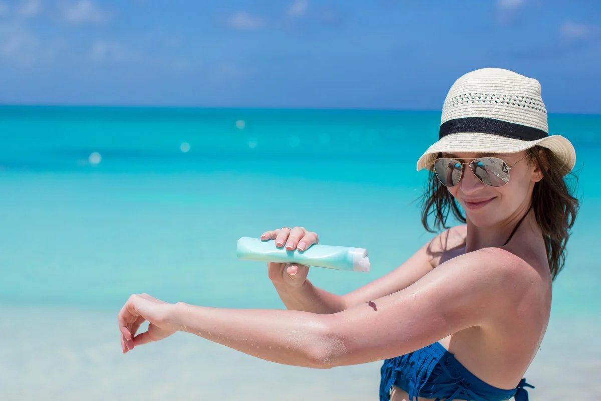 A woman in a sun hat applying sunscreen on a turquoise beach